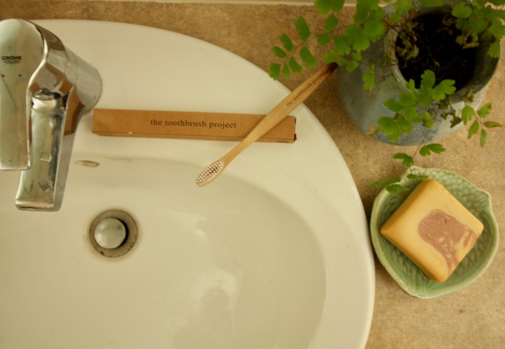 "looking down at a bathroom sink with a wooden toothbrush resting on a cardboard box with a plant and soap nearby"